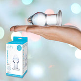 A hand is holding a clear Gläs Amethyst Rain Glass Butt Plug. The background features a soft, colorful bokeh effect. To the left of the hand and plug, there is a box with the label "Gläs" and an image of the same product on it—perfect for those exploring anal play.